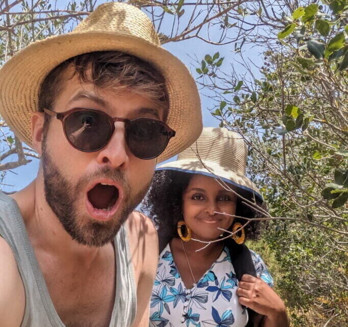 A white male and black female couple on a sunny day amidst bushes wearing straw hats
