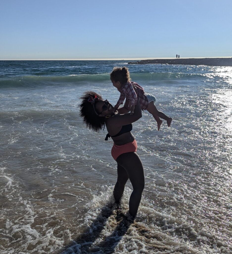 black mother with afro in a bikini with her toddler daughter in a red dress in her arms
